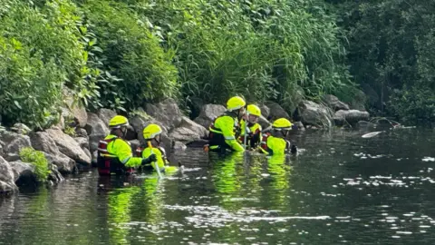 Five people in high-vis outfits and helmets waist deep in a river. Behind them to the left is the river bank, which is covered in green foliage.