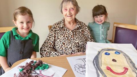 Children Eloise Pinchin (left) and Mary Shaw (right) present 78-year-old Irene Larson with artworks