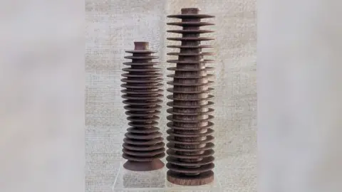 Johannes Roux Two wooden sculptures standing side by side. Both resemble wood-turned tubes and both are standing upright. They have wooden discs placed horizontally down their length. Both sit on a round wooden base.