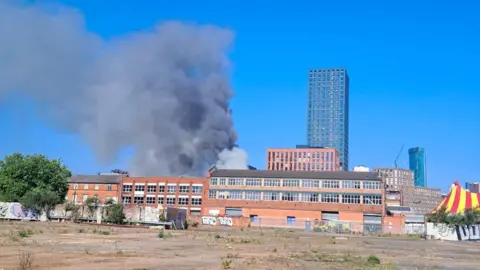 Chris Shepherd Black smoke billows into a blue sky from a factory building on an industrial estate. A high-rise tower can be seen in the background with a red and yellow tent to the right.
