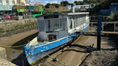 Compton Castle is a derelict boat which is stuck in the mud of Truro Harbour. Its windows are broken. Beyond it is a bus station.