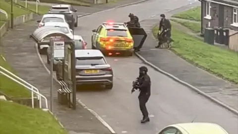 Armed police officers dressed in all black on a residential street. A police car with red lights flashing can also be seen.