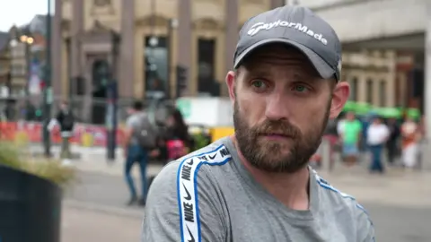 A picture of Dominic who is wearing a cap, grey top and has a brown beard, there are shoppers and buildings behind him.