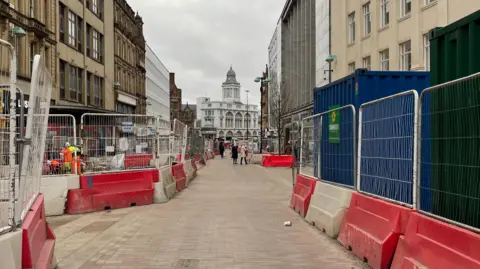 Tom MacDougall/BBC A pedestrianised street with red and white plastic barriers and fences. Large office buildings on both sides.