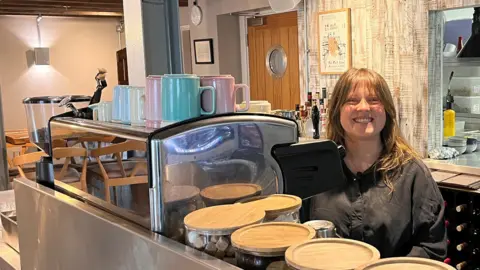 Jessica Manley, commercial director at The Yan At Broadrayne, standing being the bar at her bistro. She has long light brown hair and a fringe. She is standing behind a coffee machine with pastel coloured mugs stacked on the top. She is wearing a black shirt and is smiling broadly at the camera. 
