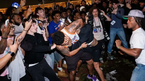 A crowd of people gather are two men fighting in the street 