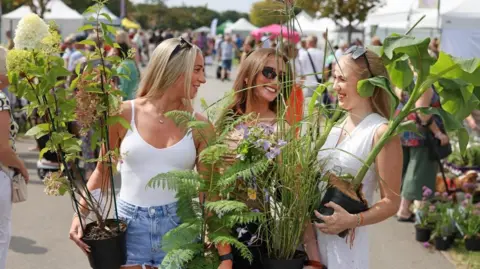 Gareth Jones Photography Three young women stand smiling at each other outside. They are each holding potted plants