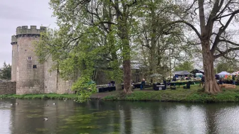 Whittington Castle stands behind the moat. To the left of the castle is a group of mature trees with trade stalls beneath. 