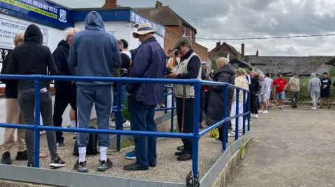 A long line of between 30 and 40 people queuing at the entrance to Southend United's ticket office, which is a small blue and white building. Outside it is a gravel car park with the queue winding through it.