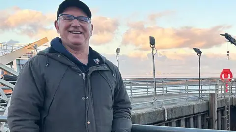 Tony Cowell standing by the sea. He is wearing a baseball hat and grey coat and has glasses and is smiling