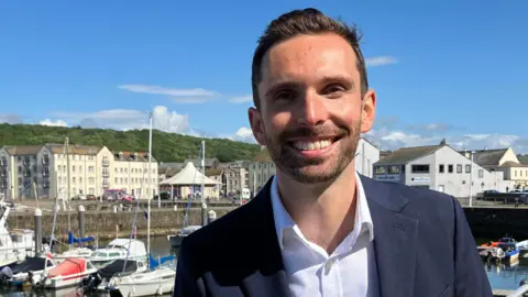 Josh MacAlister MP is standing on Whitehaven Harbour with boats and the marina behind him. He is wearing a navy blue suit and white shirt with an open collar.