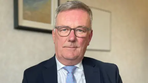 PA Media A man wearing a navy blue suit, white shirt and a pale blue tie. He is looking straight at the camera