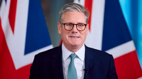 Getty Images Britain's Prime Minister Keir Starmer speaks at a press conference, during his visit to the European Commission headquarters on October 2, 2024 in Brussels, Belgium. Behind Sir Keir are two flags of the UK in the distance. He is wearing a navy suit, black spectacles and a blue tie.