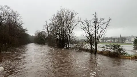 BBC A flooded road with trees and a football pitch in the background