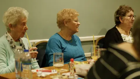 A shot from the film: Three elderly ladies are sat a table with bingo cards and daubers, drinking from beer glasses with straws. 