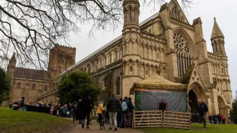 Toby Shepheard The west end of St Albans' Norman cathedral has a wooden barn outside the entrance, with many people visiting it.