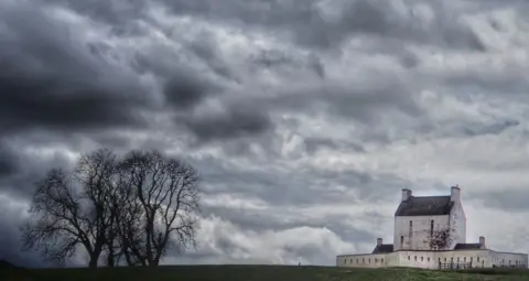 Iain Kelman A large white house surrounded by a wall with dramatic cloudy skies, and a tree in the foreground 