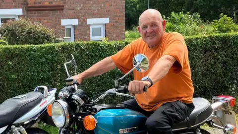 Tom Frith smiles as he poses on an old motorcycle in front of a hedge outside a garden.