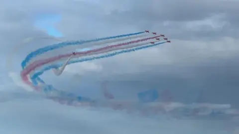 BBC Seven of the Red Arrows in formation with white, blue and red smoke coming from their tails as they fly in a curve with two aircraft weaving around the trail behind them. The sky is blue with white clouds.