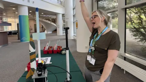 Jo Burn / BBC A woman stood in front of a model of a rocket. The woman is raising her one arm in the air and appears to be shouting in celebration.