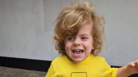 A photo of Wilfred. He is smiling and looking away from the camera. He has long, curly blonde hair and is wearing a yellow T-shirt. He is holding one arm away from his body.