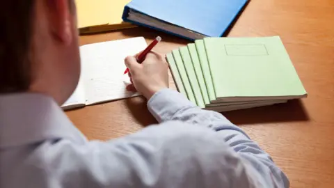 Teacher marking light green exercise books with a red pen.