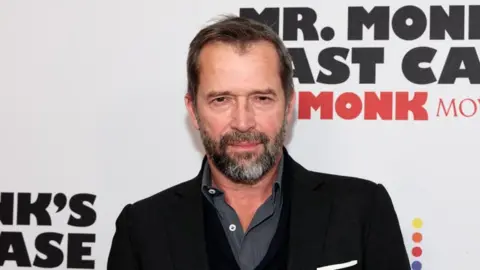 Getty Images Wearing a black jacket and dark, stripped shirt, James Purefoy gazes at the camera. He has a greying beared and stands in front of an advertising board.