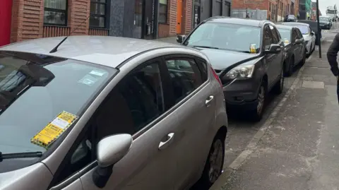 Four grey cars on a street in Birmingham's Jewellery Quarter, all of them have a grey and black Penalty Charge Notice on their windscreens.