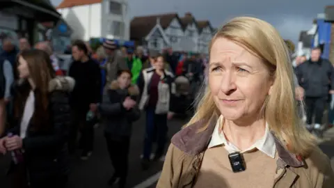 A blonde woman in a brown jacket. There is a crowd of people behind her.