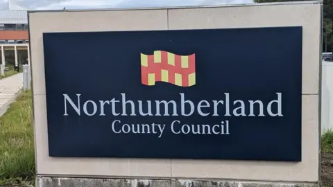 LDRS A blue sign with Northumberland County Council written on it. There is grass next to it and a path.