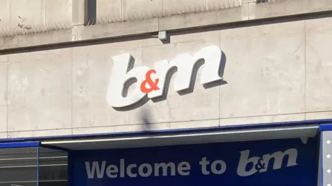 A white and orange B&M sign with signage below. On a stone wall. 