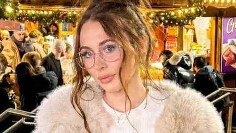 A woman with wavy brown hair which is tied up. She is wearing glasses and a fluffy beige jacket. She appears to be standing in a pub
