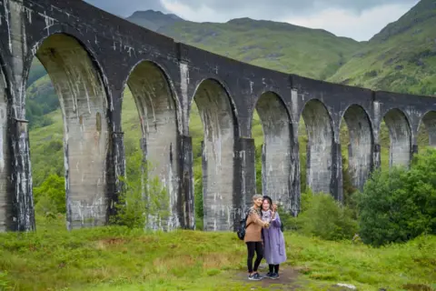 Getty Images Two tourists stand in front of the Glenfinnan Viaduct, a spectacular railway arch in the Western Highlands of Scotland. They are taking a selfie with a selfie stick.