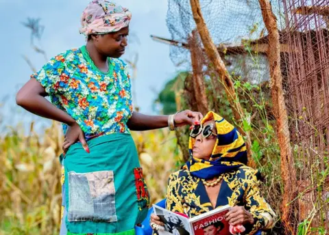 Luxury Media Zambia Diana Kaumba stands outside in traditional Zambian clothing looking down at her grandmother who is sitting in a navy and gold patterned jacket, scarf and sunglasses - holding a book titled Hollywood Fashion, with Audrey Hepburn on the cover.