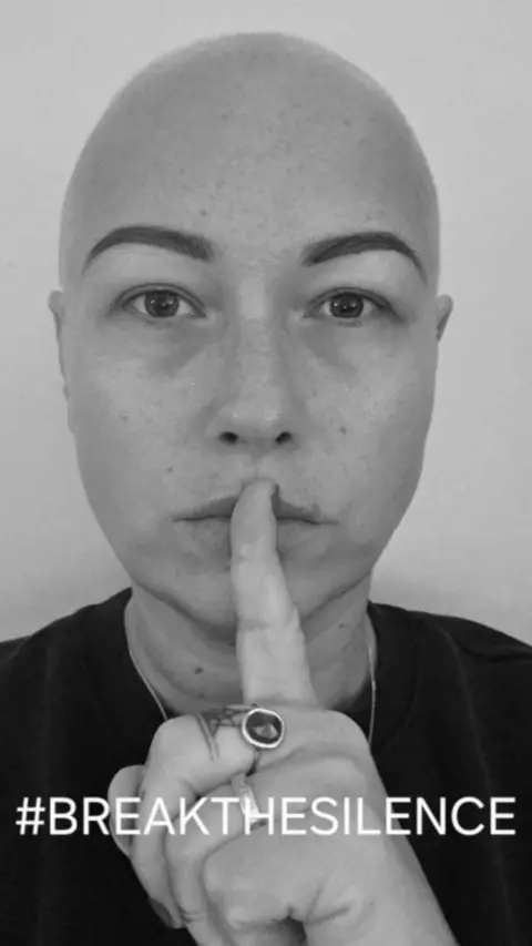 A black and white image of a woman with no hair holding her finger to her lips.
