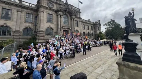 A crowd of people outside South Shields Town Hall, a large stone building in the Edwardian Baroque style. There are columns on either side of the doorway, which is topped by a large pediment and a flag pole. The ground floor features a row of arched windows, and the upper floor square ones. The crowd, standing on the steps leading towards the entrance, and round to the side, are holding flags and banners.