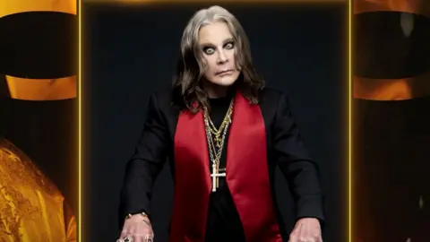 A photo of Ozzy Osbourne wearing a black jacket with large red lapels and several gold chains with crosses around his neck. He looks intensely at the camera.