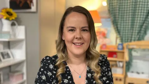 Emma has long brown curly hair and is pictured in a school classroom. She is wearing a black patterned dress.