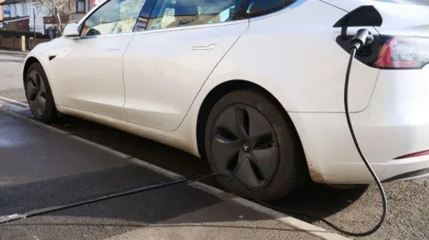 Oxfordshire County Council A white EV has a charging cable struck in it. The cable itself is running through a small, narrow trench in the path.