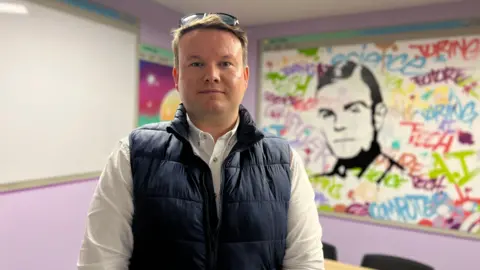 Sebastian looks at the camera as he wears a navy blue gillet and a white shirt. He has short hair with sunglasses on the top of his head. Behind him is a conference table and chairs and on the wall there is a white board with artwork on it.
