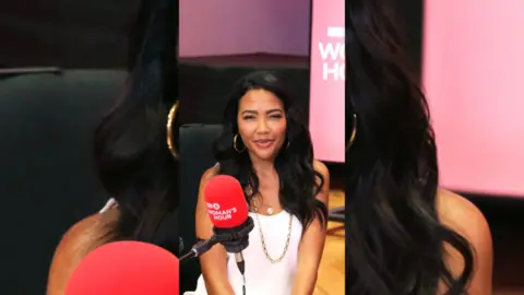 Emma Grede has long dark hair and is wearing a white top. She's sat in front of a red microphone.