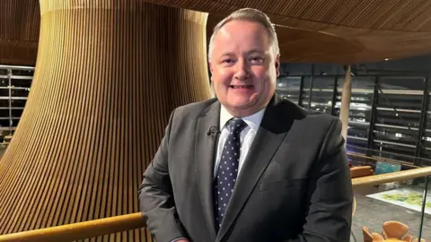 Darren Millar is stood slightly to the right of the middle of the shot, on a balcony overlooking the funnel in the middle of the Senedd building. He is in a suit, tie and white shirt, and is smiling.