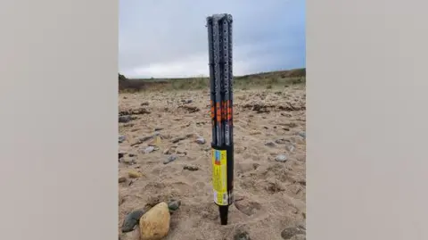 Sharon Bell A large firework canister. The black plastic tubing is long and tall. Its pointed base is stuck in the sand. It looks like it can hold several fireworks. Rocks are also strewn among the sand.