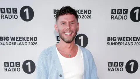 Getty Images Dean McCullough wears a white tshirt and blue cardigan. He stands in front of a board that says BBC Radio 1 Sunderland 2026.