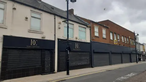 LDRS Street with former retail units, the first two on the right have white upper storeys, while the one of the left is brick. All have closed black shutters covering both the downstairs windows and doorways.