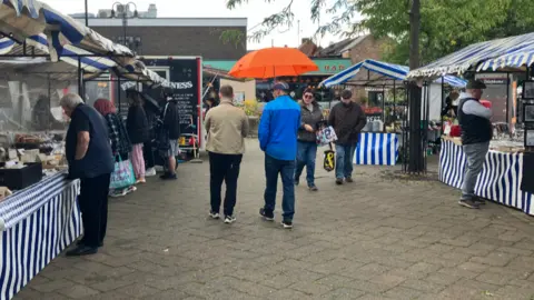 Blue and white-striped market stalls either side of a paved area with people walking towards and away 