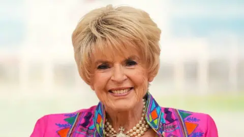 PA Media Gloria has short blonde hair and is wearing a bright patterned jacket with a pearl necklace.