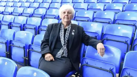 Lily Barnes is photographed sitting in one of Goodison Park's stands and smiling at the camera. There are rows of blue seats  all around her.