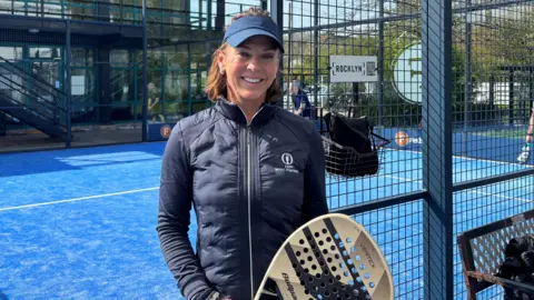 Deborah is standing in-front of a Padel court holding a gold and black racquet. The court is blue with a black fence. There is a building in the back ground. 