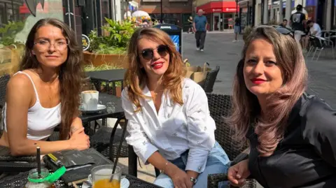 Contributed Three people sitting at an outdoor cafe table on a pedestrian shopping street. The table holds drinks, a partially eaten dessert, and personal items. Shops, cafe seating, and pedestrians line the street behind them in bright afternoon sunlight.